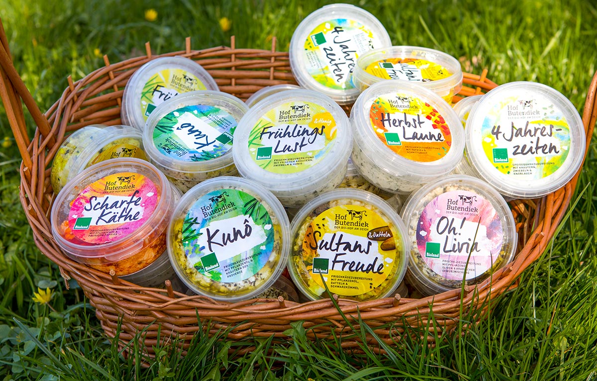 unsere-produkte-auswahl-im-korb Zu sehen ist ein Weidenkorb, im Gras zwischen Butterblumen abgestellt. Dieser ist gefüllt mit unseren unterschiedlichen Frischkäsebechern.